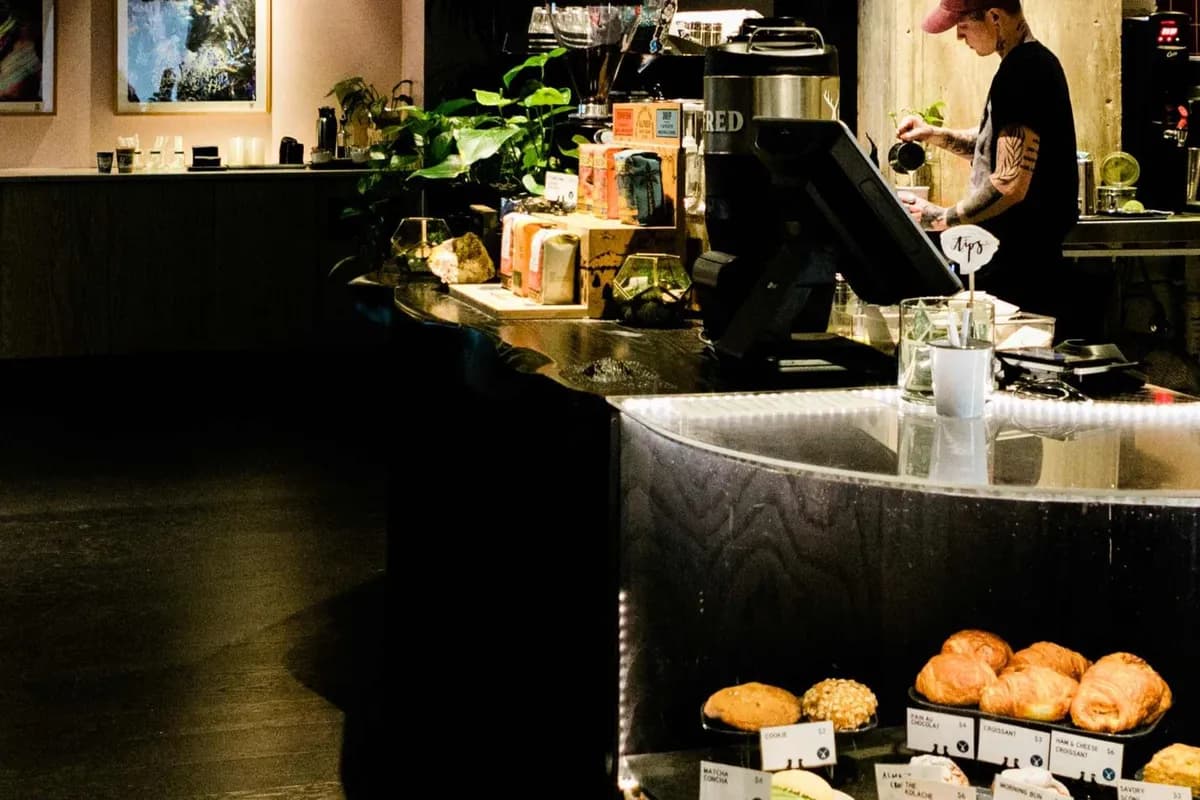 Barista pours milk behind the curved black counter at Alfred Coffee, with pastries in the case and plants and framed art behind