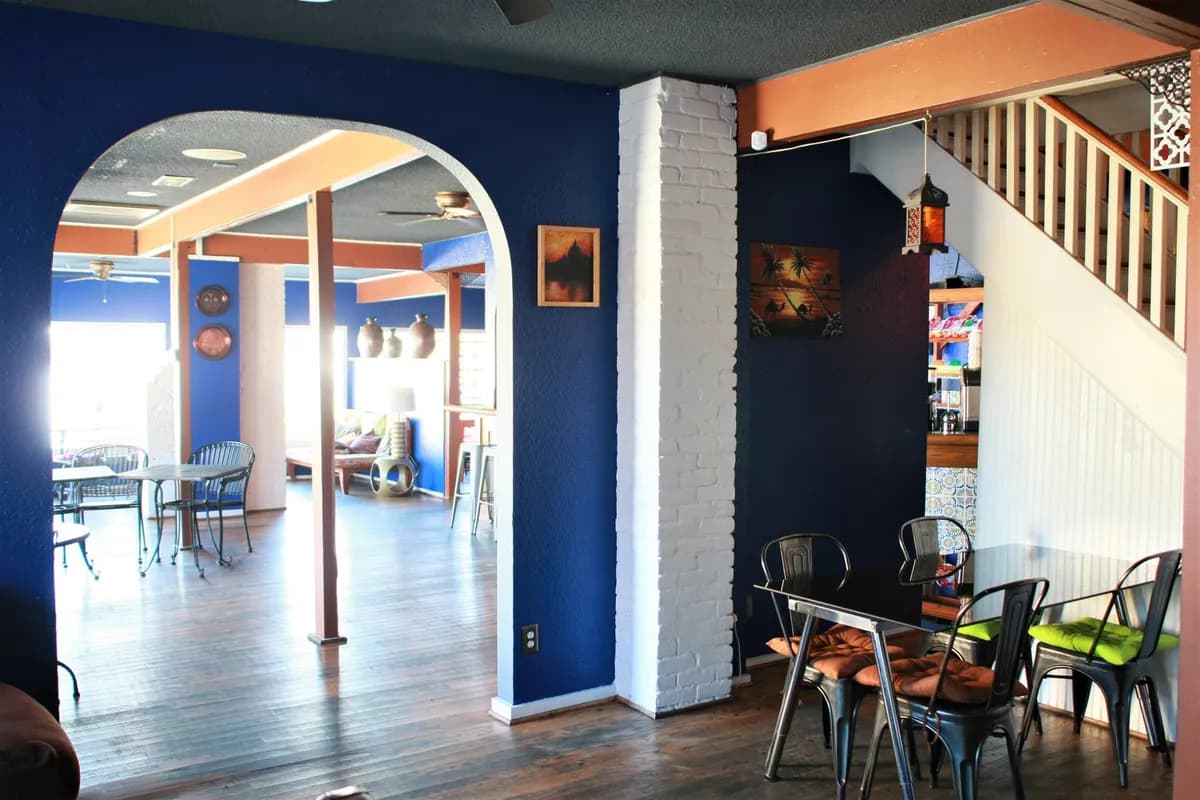 Arched doorway into a bright blue-walled lounge with wood floors, cafe tables and a staircase