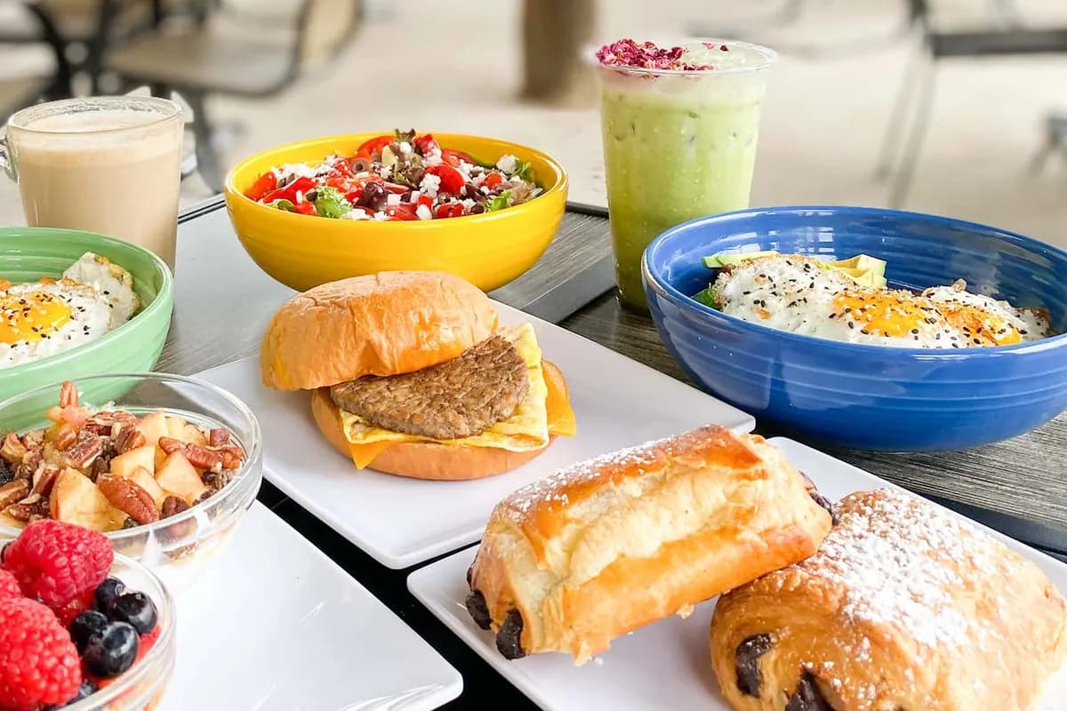 Patio table spread of colorful breakfast bowls, sandwich, pastries and iced drinks