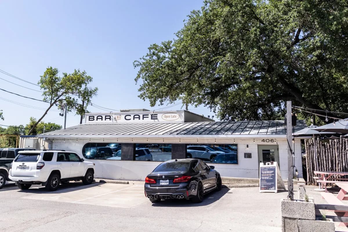 Low white bar-and-cafe building with metal roof, parked cars out front and a large oak tree behind