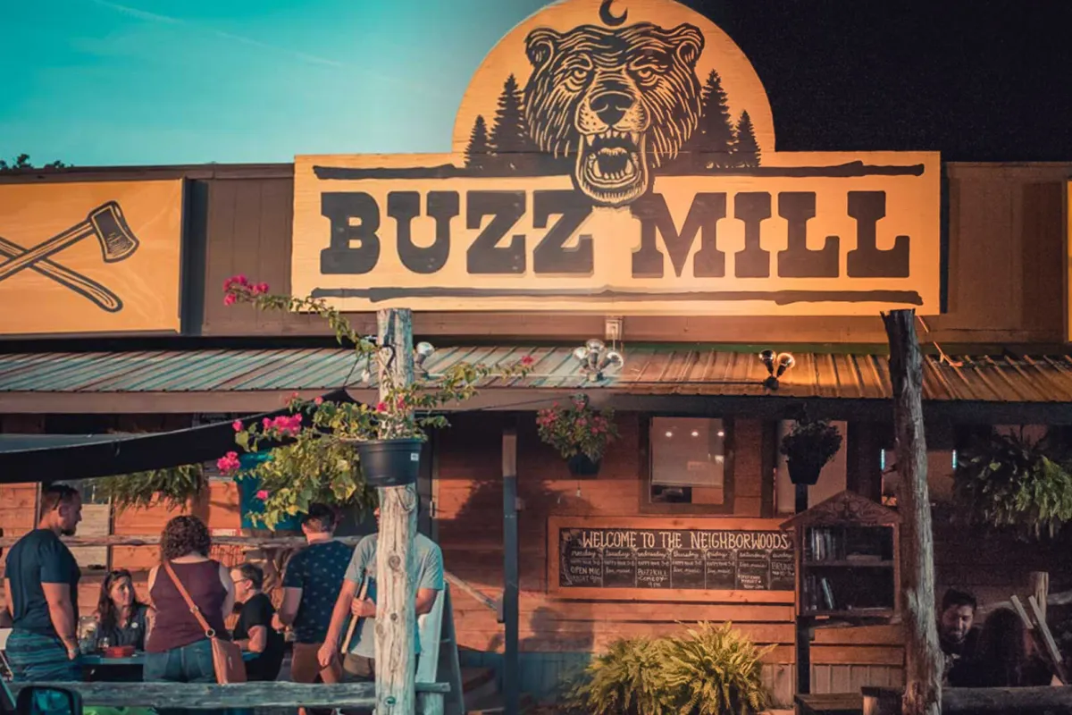 Buzz Mill's glowing bear-and-axes sign over a rustic cabin-style patio at dusk with people gathered