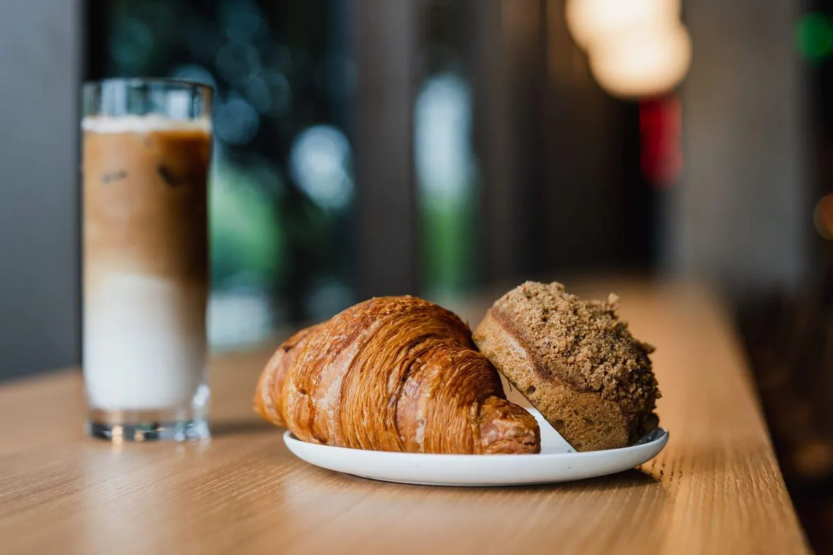 Iced latte beside a croissant and chocolate pastry on a plate on a wooden counter