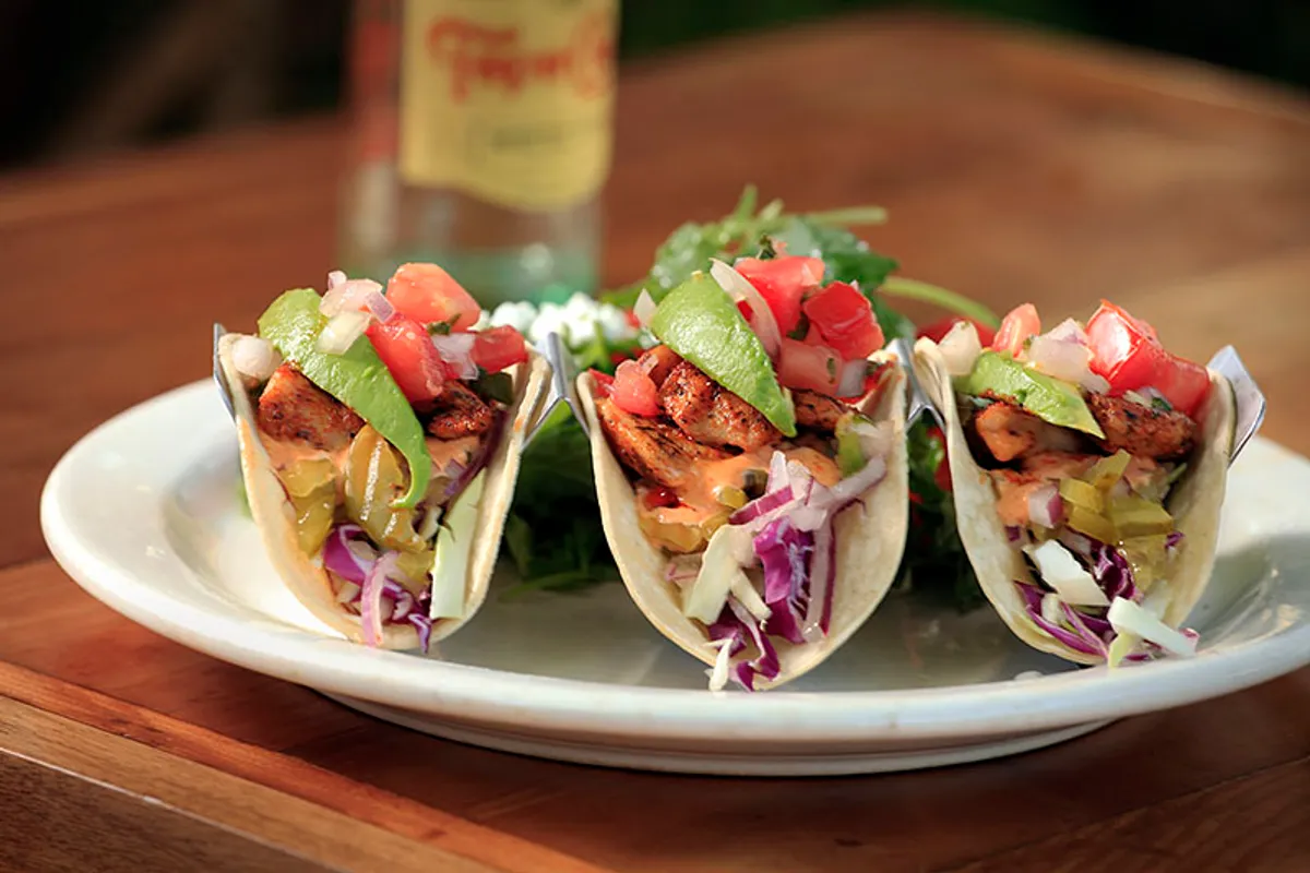Plate of three fish tacos with avocado, pico and slaw on a wooden table beside a bottled drink