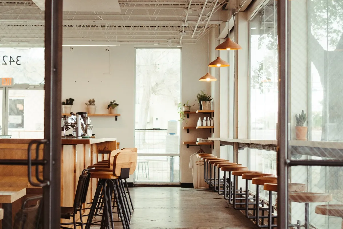 Sunlit cafe with a wood counter, a row of tall stools along the window bar, and copper pendant lamps