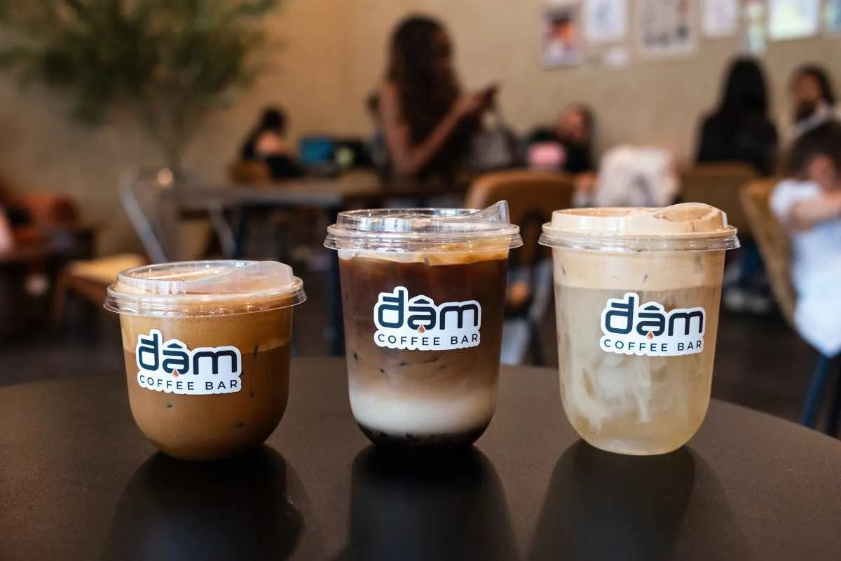 Three iced coffees with Đậm Coffee Bar stickers on a table, customers seated in the cafe behind
