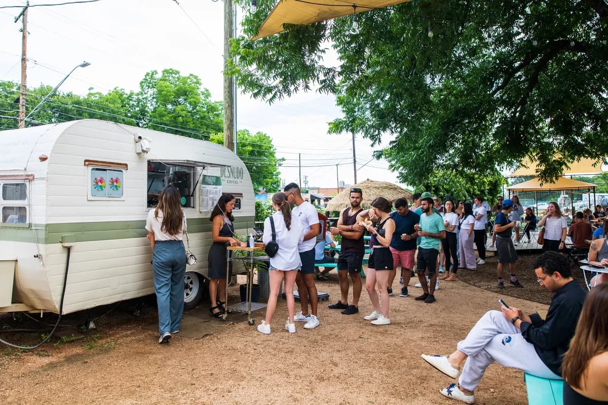 Customers queue at the Desnudo Coffee vintage trailer window under a large shade tree in the yard