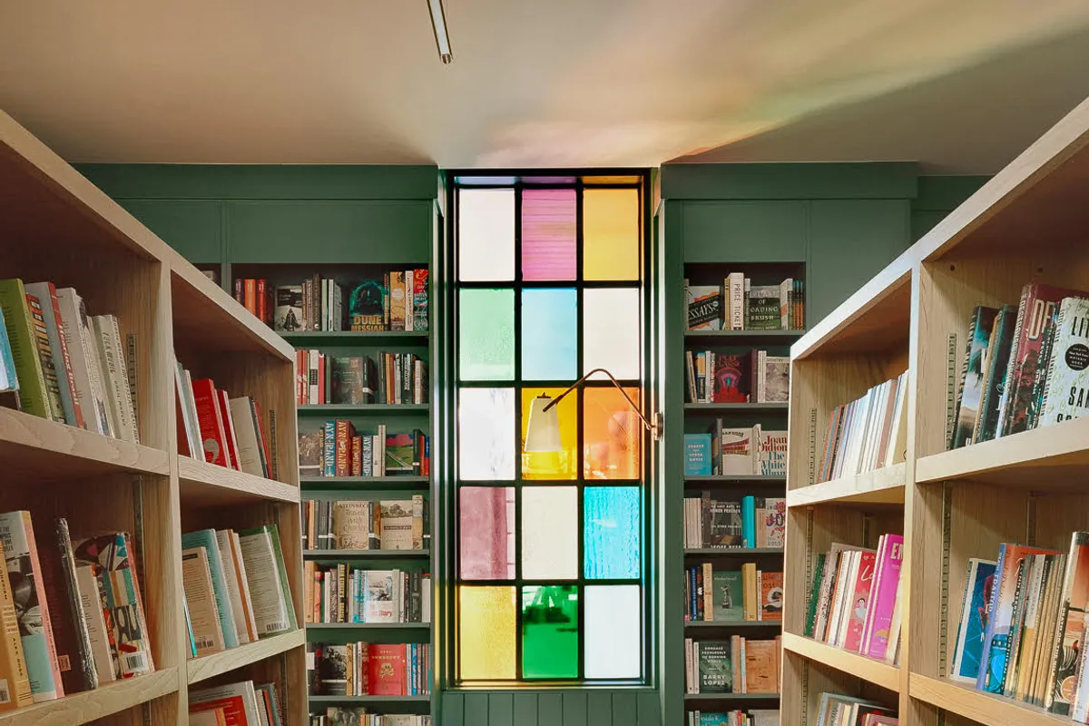 Bookstore aisle between wooden shelves leading to a green nook with a colorful stained-glass window and bench