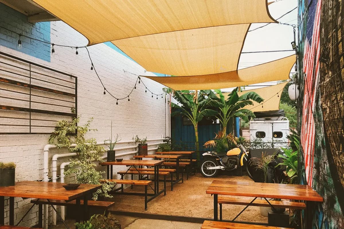 Covered outdoor patio with picnic tables, string lights, banana plants and a vintage yellow dirt bike against a mural wall