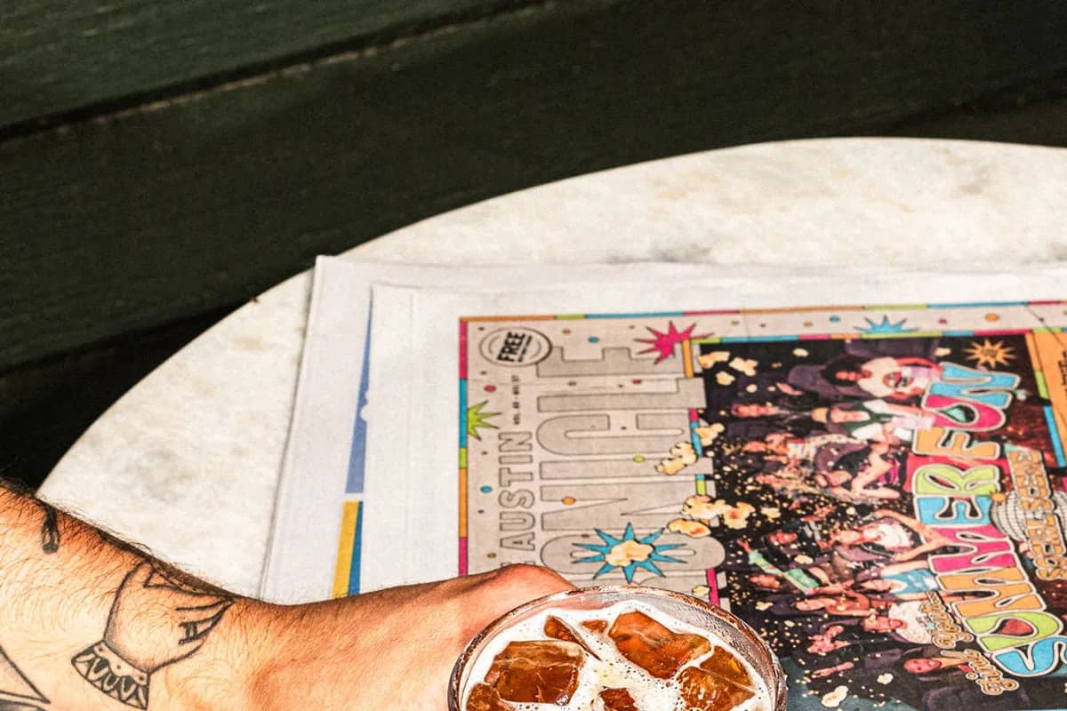 Hand holding an iced drink in a faceted glass on a marble table beside an Austin Chronicle