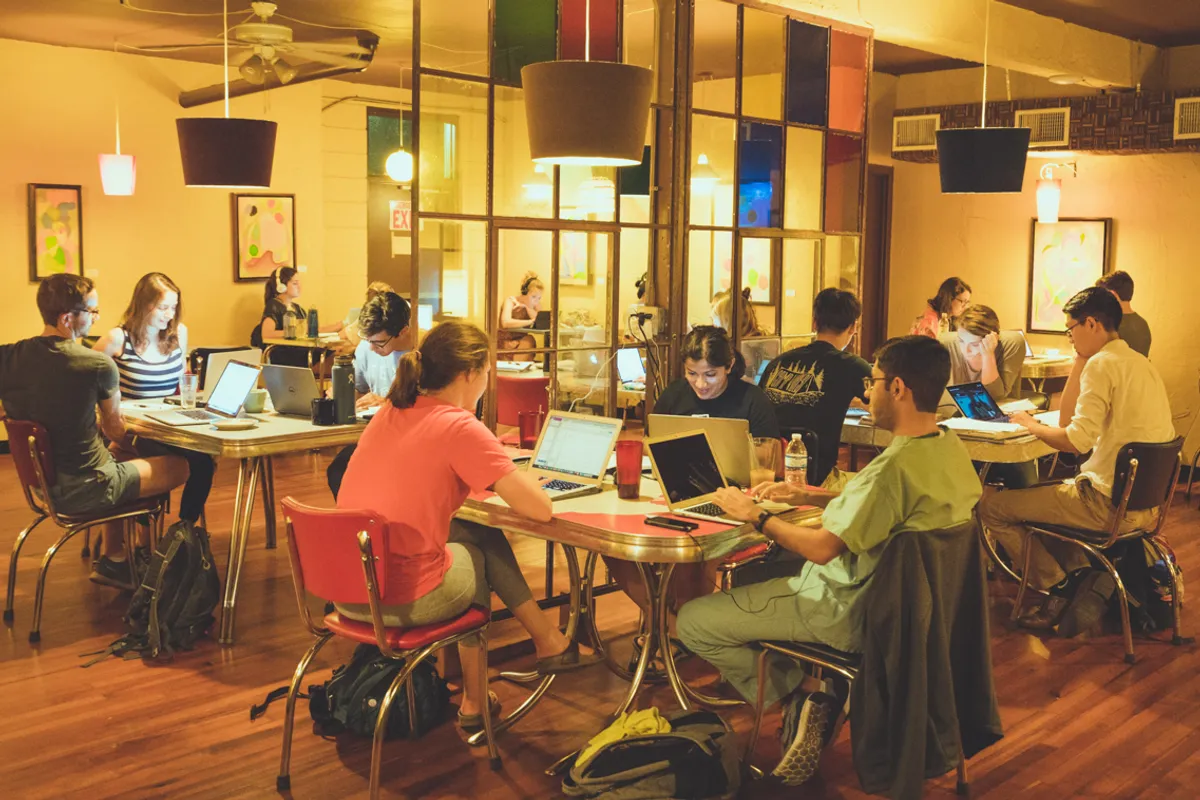 Students working on laptops at retro tables with red chairs inside a warmly lit coffeehouse