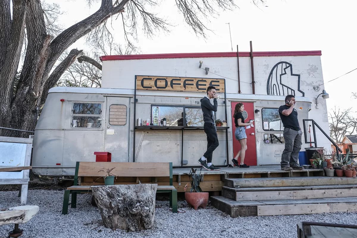 Vintage trailer coffee stand with a COFFEE sign and three people chatting on the deck beside a gravel yard