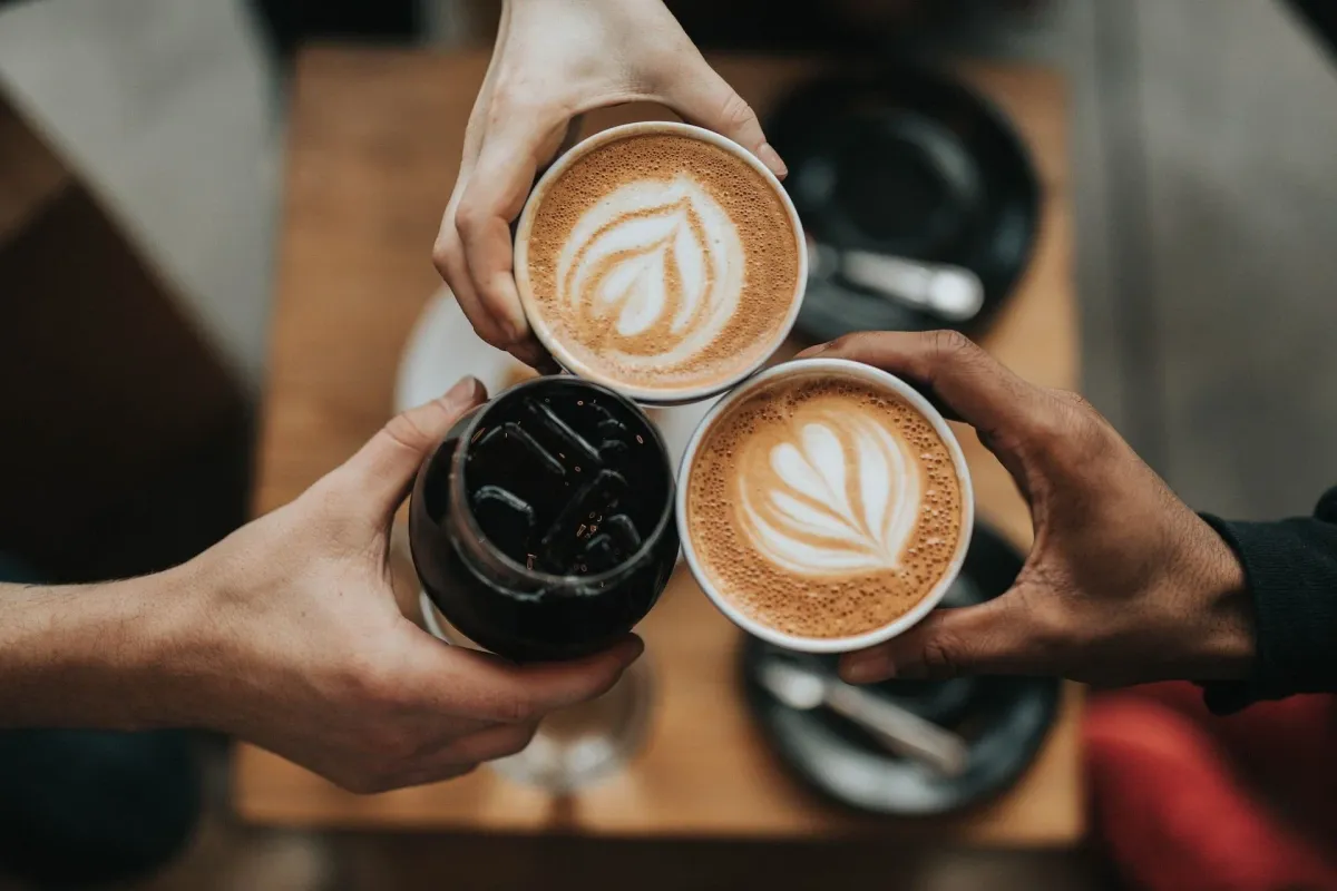Three hands cheers-ing two lattes with art and an iced coffee over a wooden table