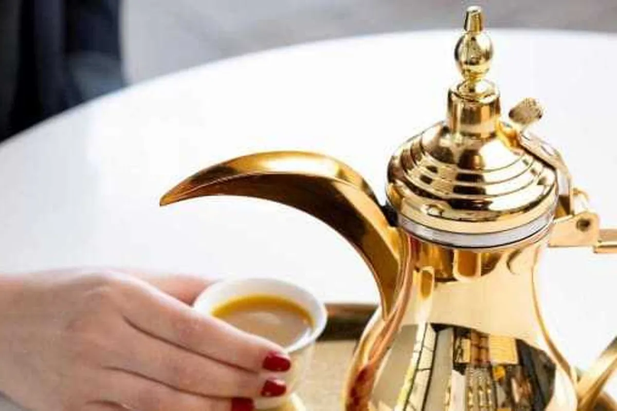 Hand taking a small cup of Yemeni coffee beside a gold dallah coffee pot on a white table