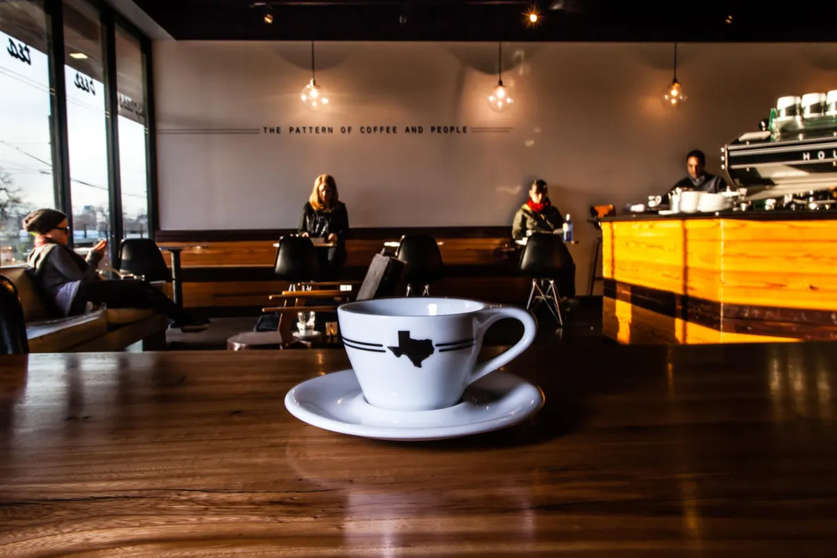 Customers seated along a window wall beneath a lettered wall slogan, with espresso bar and pendant lamps at right