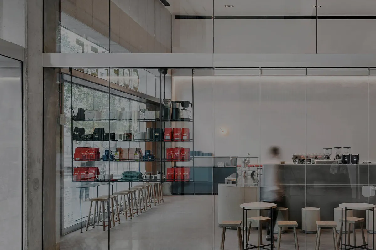Glass-fronted cafe with shelves of red coffee bags, a slim espresso bar, and small wooden stools