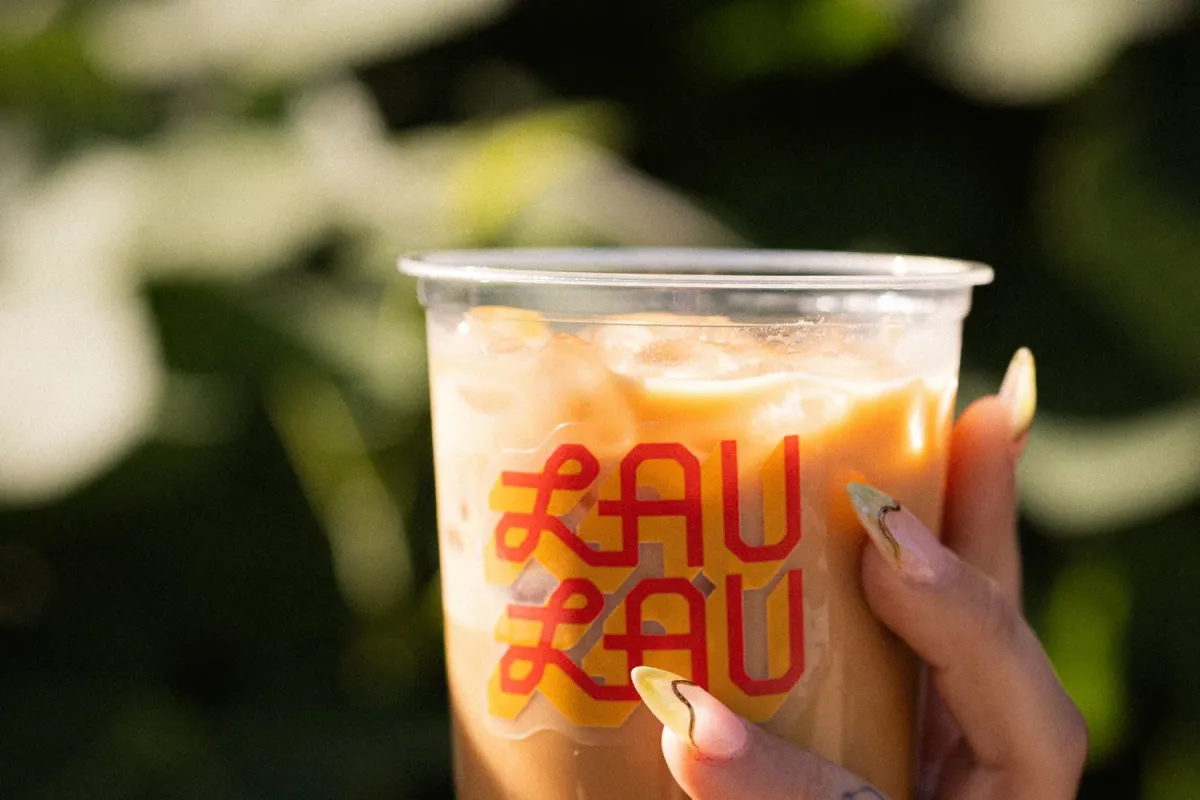 Hand holding an iced coffee in a clear Lau Lau branded cup against sunlit foliage