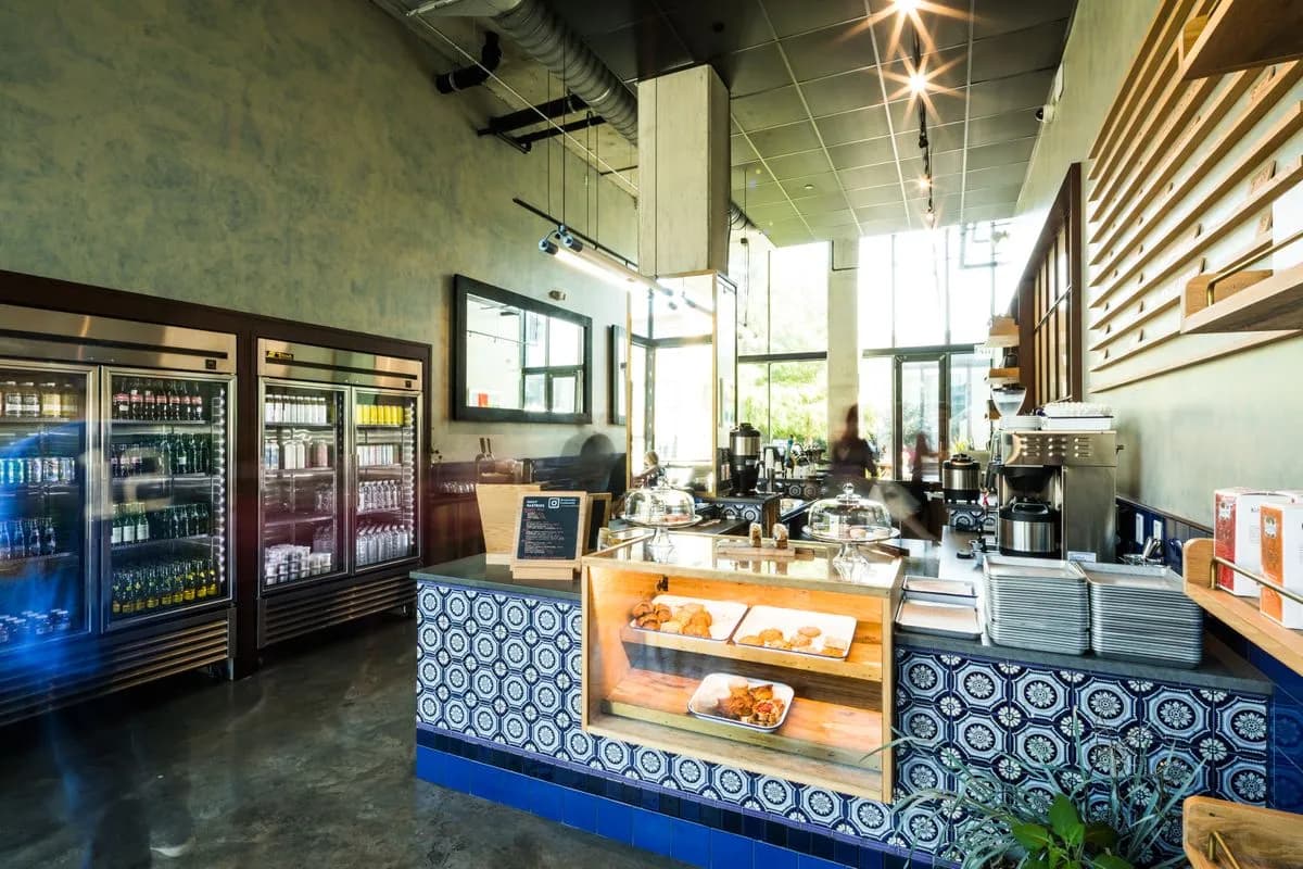 Bright cafe interior with tiled blue pastry counter, glass-door drink coolers and large windows