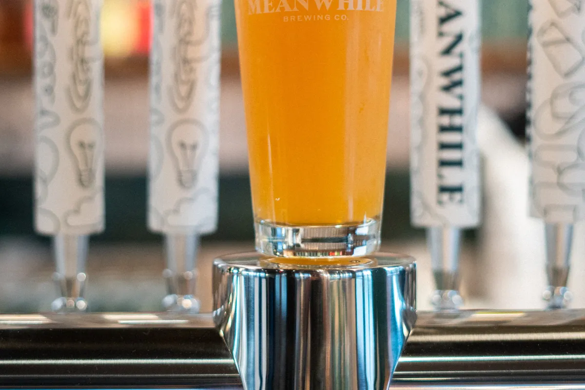 Pint of golden beer in a Meanwhile glass in front of a row of white tap handles at the bar