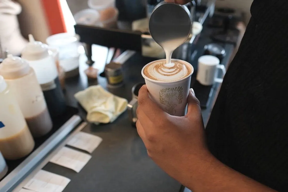 Barista pouring steamed milk into a branded paper cup of latte at the counter