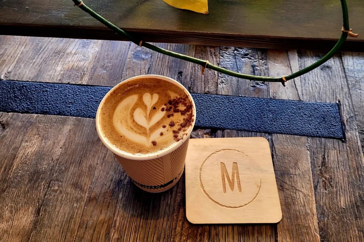 Latte with leaf art in a paper cup beside a wooden M coaster on a reclaimed wood table