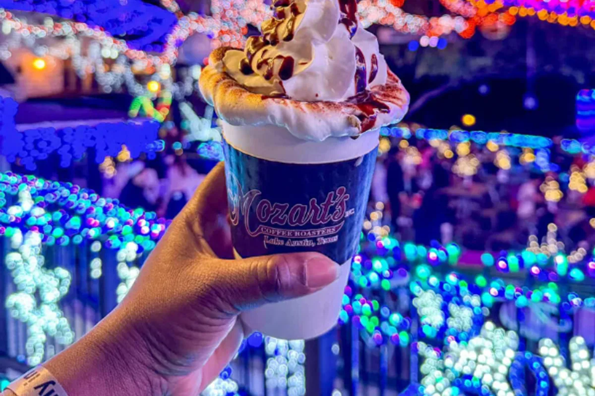 Hand holding a Mozart's-branded cup of whipped hot chocolate in front of holiday lights on the lakeside patio
