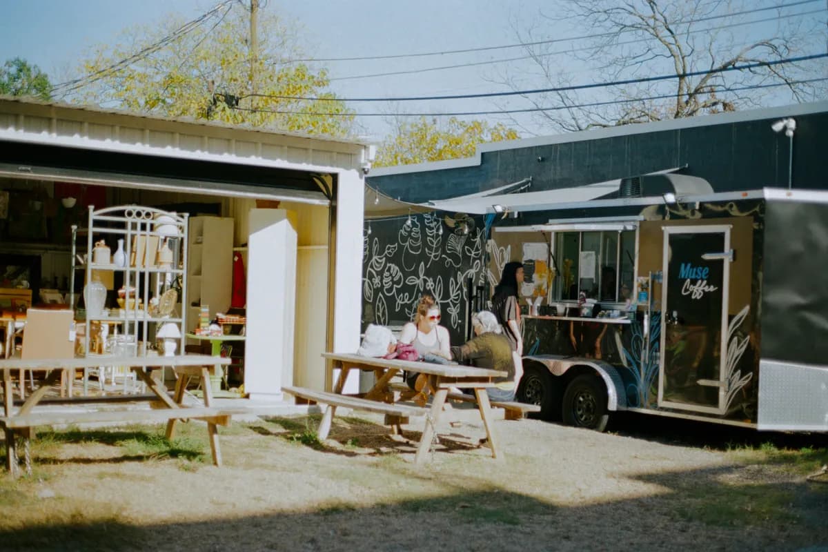 Guests sit at picnic tables beside the black Muse Coffee trailer, with a chalkboard mural and vintage shop beyond