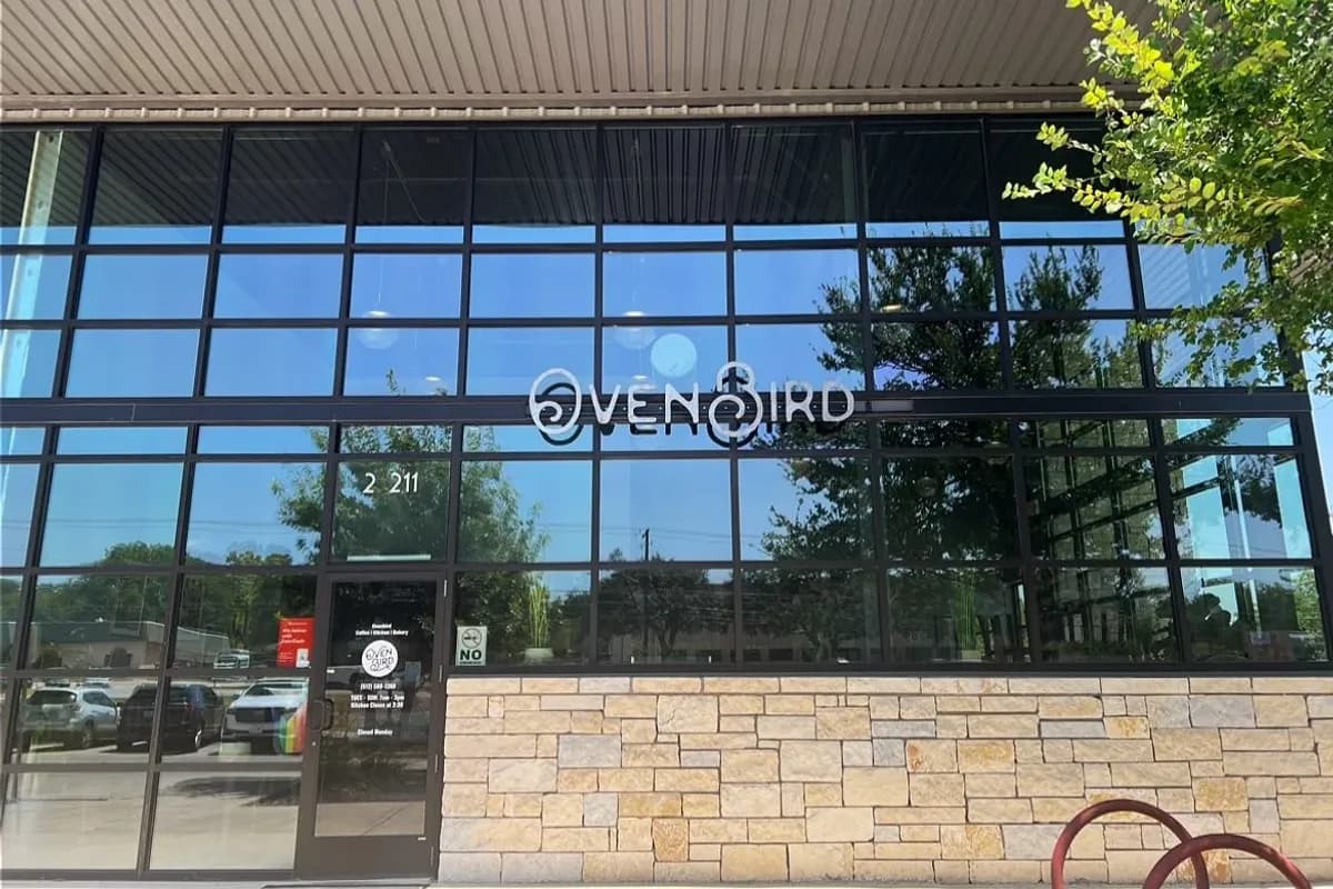 Ovenbird storefront with floor-to-ceiling black-framed windows, stone base and lettered sign under a metal canopy
