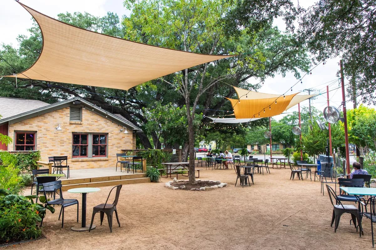 Gravel beer garden at Radio Coffee and Beer with shade sails, live oaks, small stone building and scattered tables