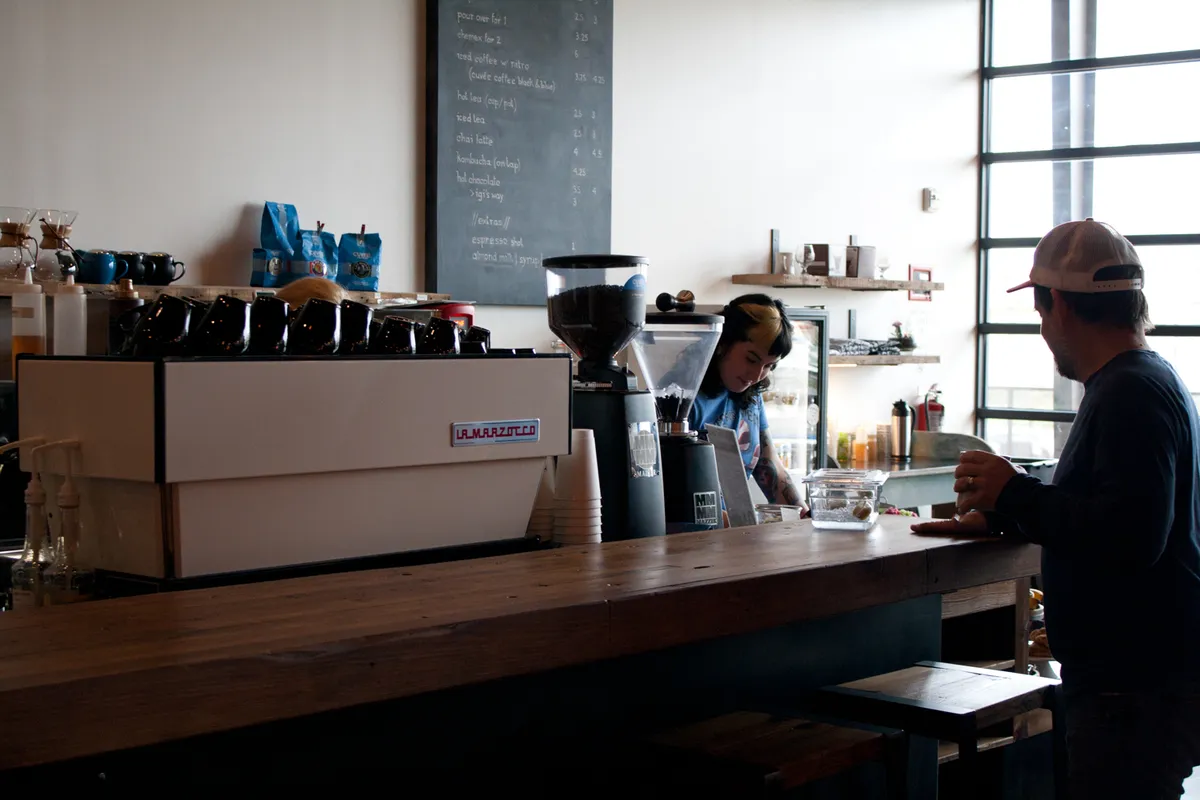Stouthaus counter with espresso machine and grinder, chalkboard menu, a barista and a customer at the wooden bar