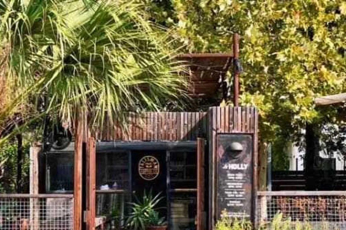 Small wood-slat container coffee bar with Talisman logo and La Holly menu board, framed by palms and trees