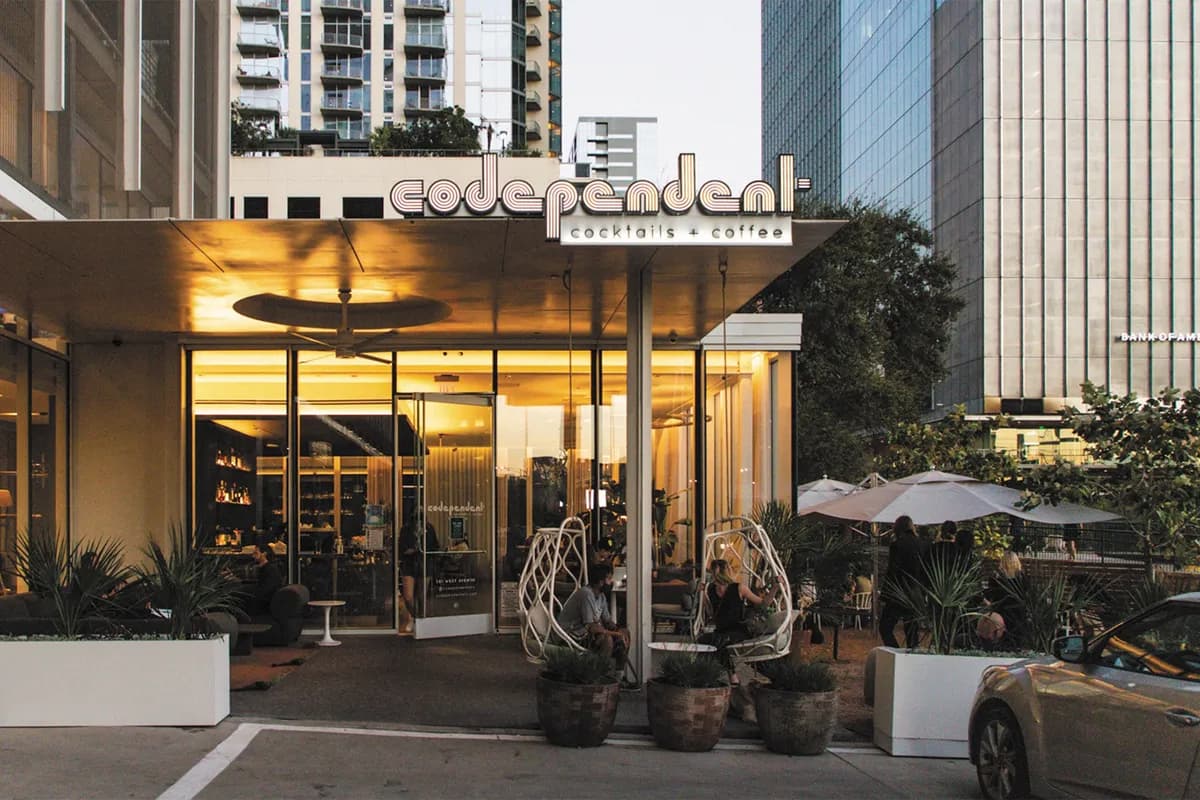 Glass-fronted Codependent storefront glowing at dusk with patio umbrellas and downtown towers behind